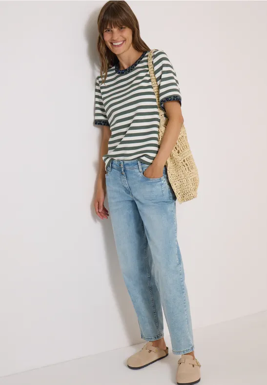 Frau mit gestreiftem Shirt, Jeans und Strohtasche lehnt an weißer Wand und lächelt.
