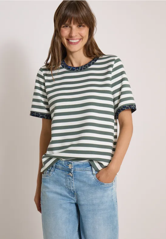 Eine lächelnde junge Frau in einem gestreiften T-Shirt und Jeans, eine Hand in der Hosentasche vor hellem Hintergrund.