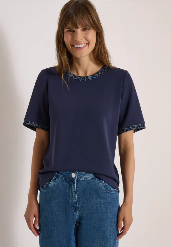 Frau in dunkelblauem T-Shirt mit gemustertem Saum und blauer Jeans, lächelnd