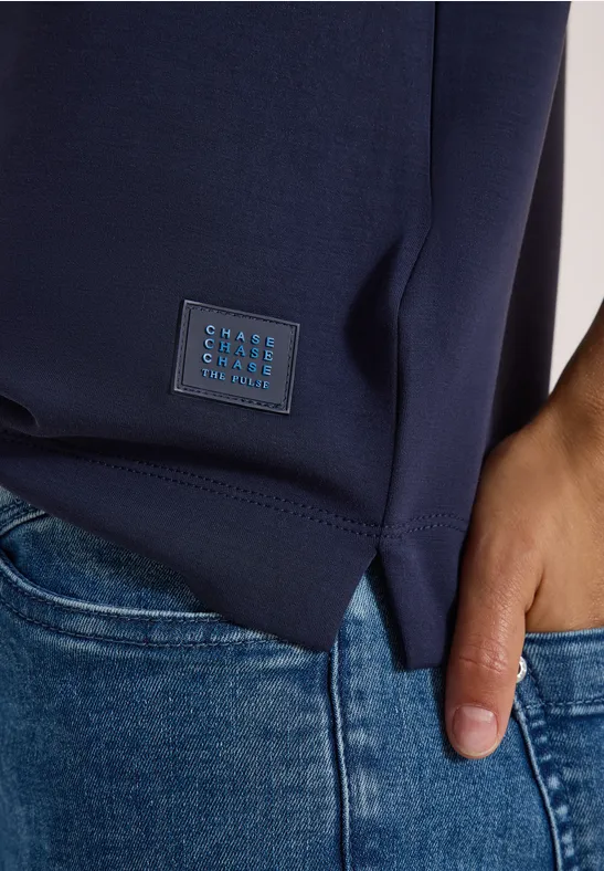 Ein dunkelblaues Shirt mit quadratischem Label am Saum und blaue Jeans, Hand in der Tasche.