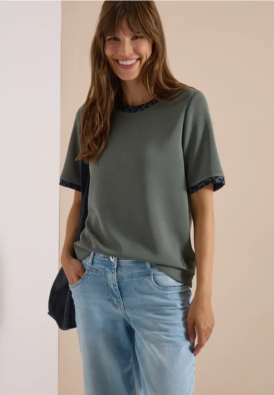 Eine lächelnde Frau trägt ein grünes Shirt und blaue Jeans.