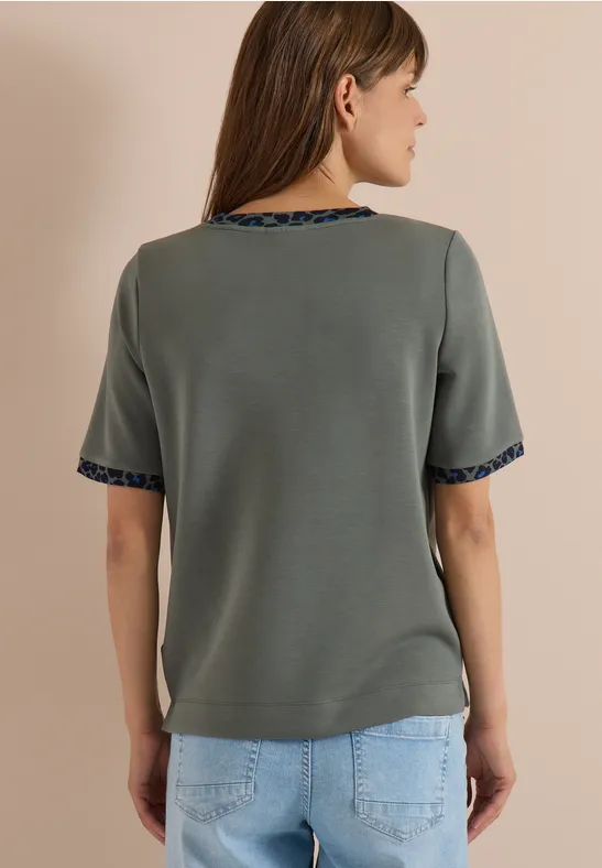 Frau steht mit dem Rücken zur Kamera, trägt graues Kurzarmshirt mit gemustertem Saum am Arm- und Halsausschnitt.
