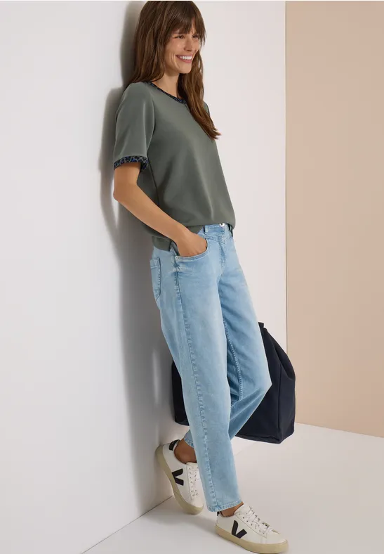 Junge Frau mit grünem Shirt, hellblauen Jeans und Sneakern lehnt an Wand, lächelt und trägt Tasche.