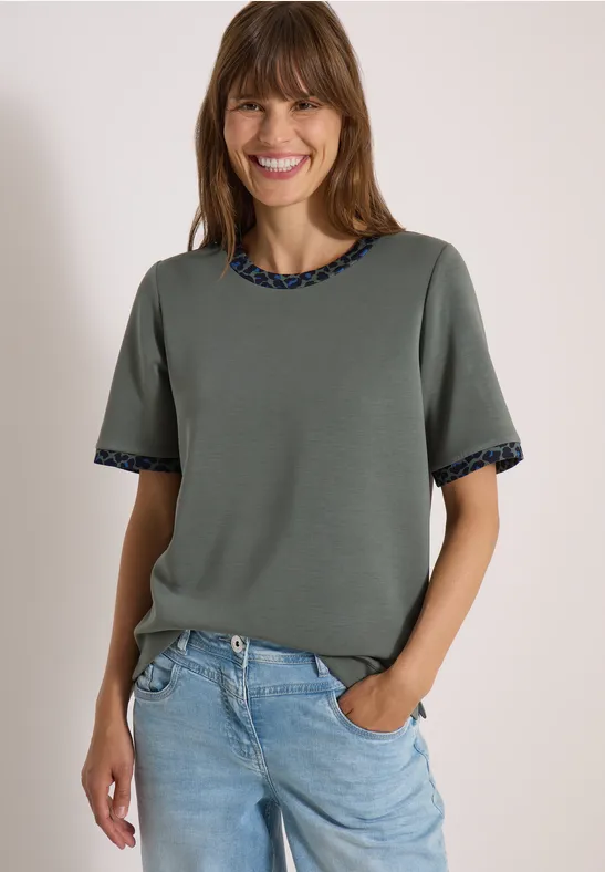Lächelnde Frau in khakifarbenem kurzärmeligen Oberteil mit gemusterten Abschlüssen und hellblauen Jeans.