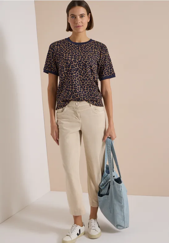Eine Frau trägt ein T-Shirt mit Leopardenmuster, eine helle Chinohose, weiße Sneaker und hält eine blaue Stofftasche.