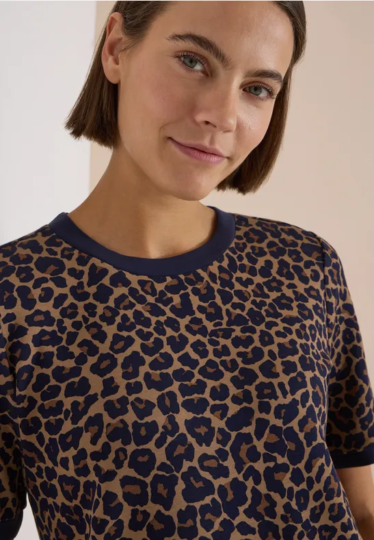 Frau mit kurzen braunen Haaren trägt ein braun-blaues Leoprint-T-Shirt und blickt freundlich zur Kamera.