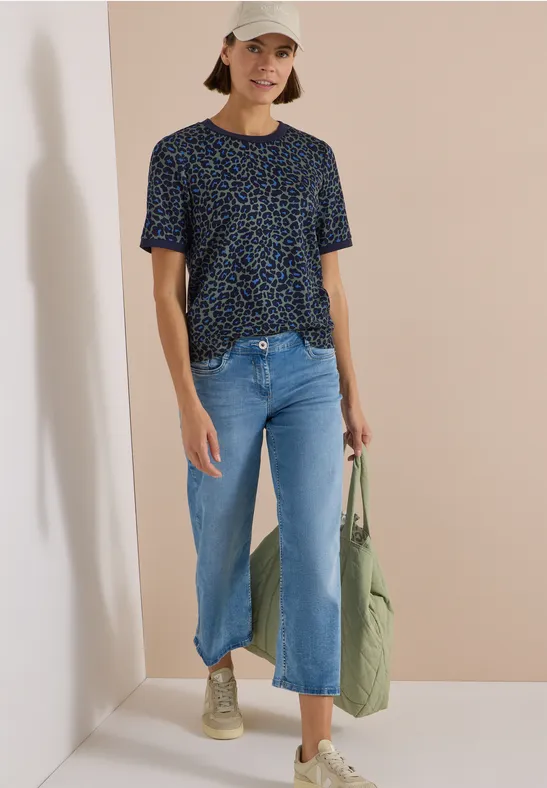 Frau in lässigem Outfit: Gemustertes T-Shirt, weite Jeans, beige Sneaker und Cap, mit grünem Stoffbeutel.