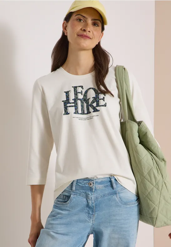 Vrouw in denim, wit 3/4-shirt met ‘HERO HIKE’-opdruk, gele pet en groene doorgestikte tas.