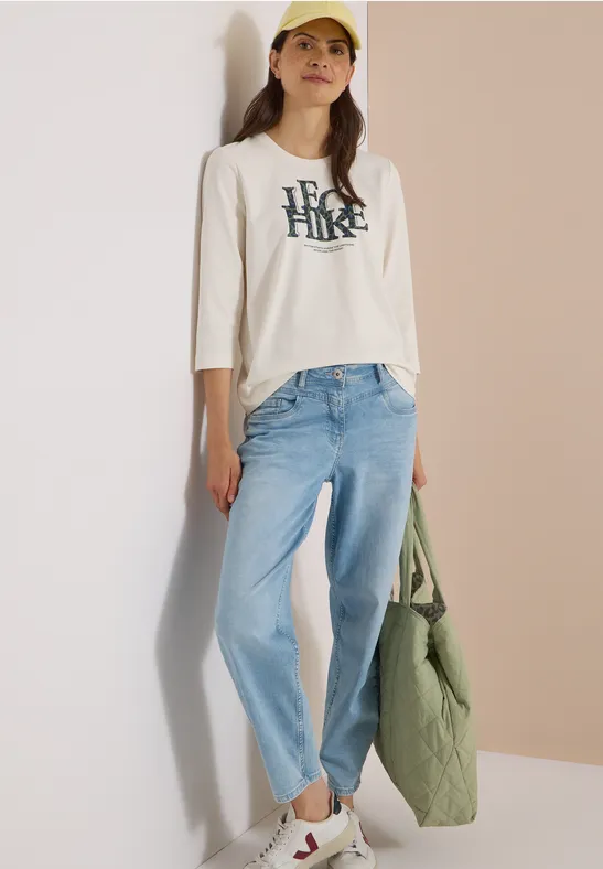 Jonge vrouw in wit shirt, lichtblauwe jeans en gele pet, met een groene tas.