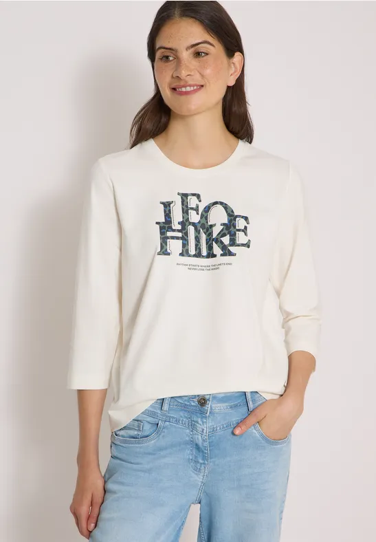 Jonge vrouw draagt crème sweat met "ECO HIKE"-print, driekwartmouwen en lichtblauwe jeans.