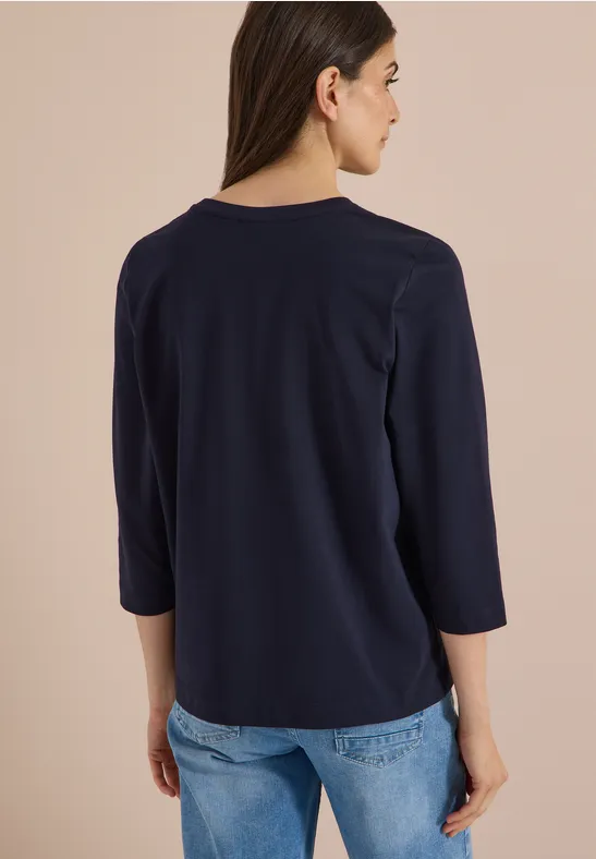 Rugaanzicht van een vrouw in een donkerblauwe driekwart mouw top en lichtblauwe spijkerbroek.