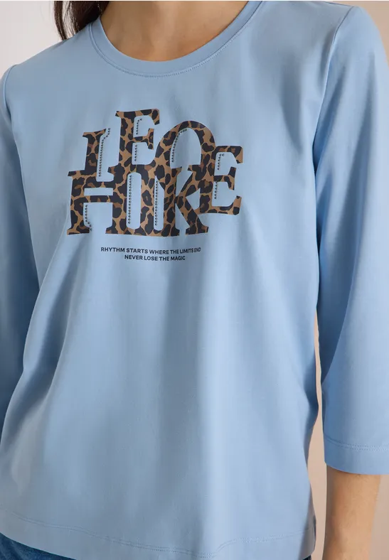 Close-up van een lichtblauw shirt met driekwart mouwen en opvallende 'LEO HIKE'-tekst in luipaardprint.