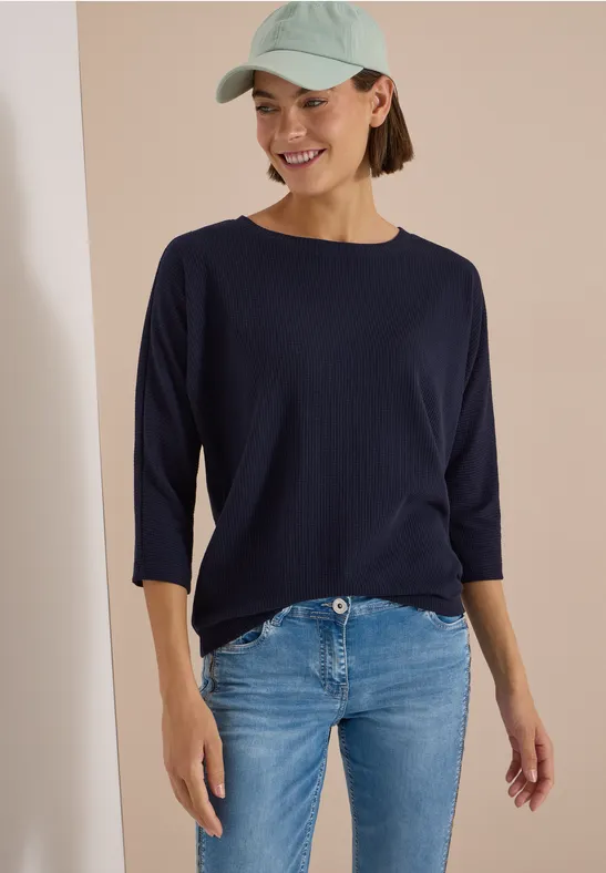 Frau trägt marineblaues 3/4-Arm-Shirt, hellblaue Jeans und eine hellgrüne Baseballcap, lächelnd vor hellem Hintergrund.
