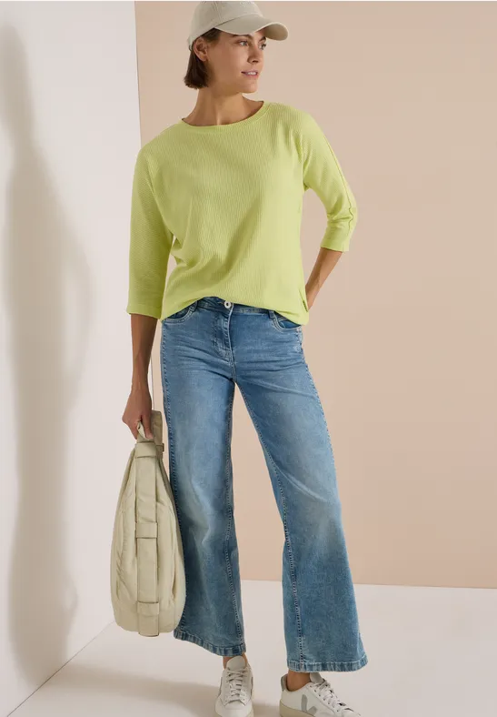 Eine Frau lehnt an der Wand, trägt hellgrünen Pullover, Jeans, Cap und Tasche.
