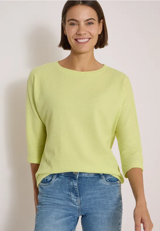Eine lächelnde Frau trägt einen hellgrünen Pullover mit 3/4-Ärmeln und blaue Jeans vor neutralem Hintergrund.