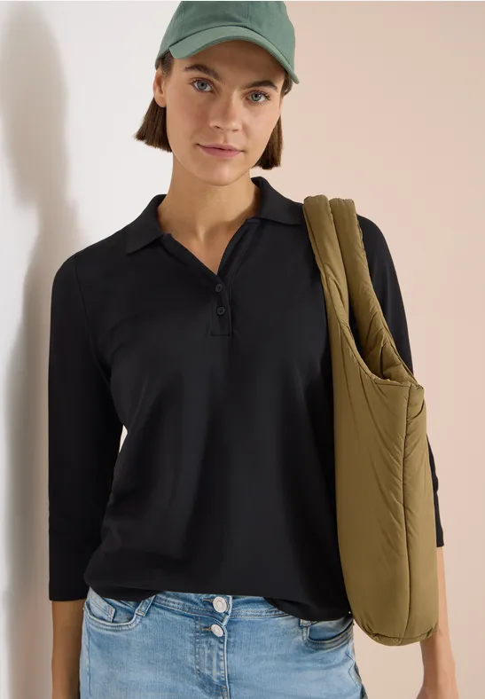 Frau trägt schwarzen Poloshirt, grüne Mütze und olivgrüne, gesteppte Umhängetasche