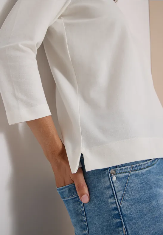 Frau trägt weißes Shirt mit 3/4-Ärmeln und blaue Jeans, Hand in Hosentasche.