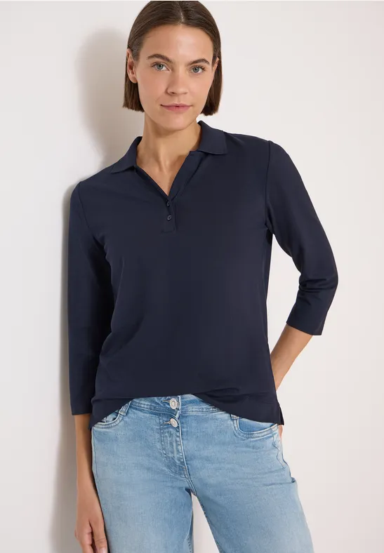 Frau trägt ein dunkles Poloshirt mit 3/4-Ärmeln und helle Jeans vor weißem Hintergrund