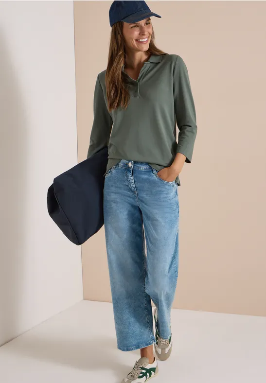 Frau mit blauer Jeans, olivgrünem Shirt und Cap hält navy Jacke, lässiger Alltagslook.