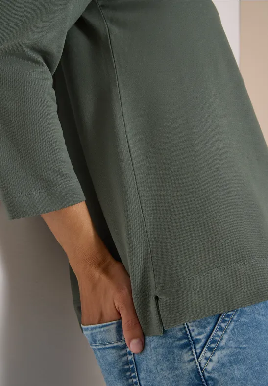 Person lehnt an Wand, trägt grünes Shirt mit 3/4-Ärmeln und blaue Jeans, Hand steckt in Tasche.