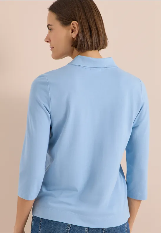 Frau mit dunklem Bob trägt hellblaues 3/4-Arm-Shirt, steht blickabgewandt vor neutralem Hintergrund.