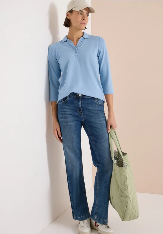 Frau trägt hellblaues Poloshirt, weite Jeans und hält eine grüne Stofftasche vor zweifarbigem Hintergrund.