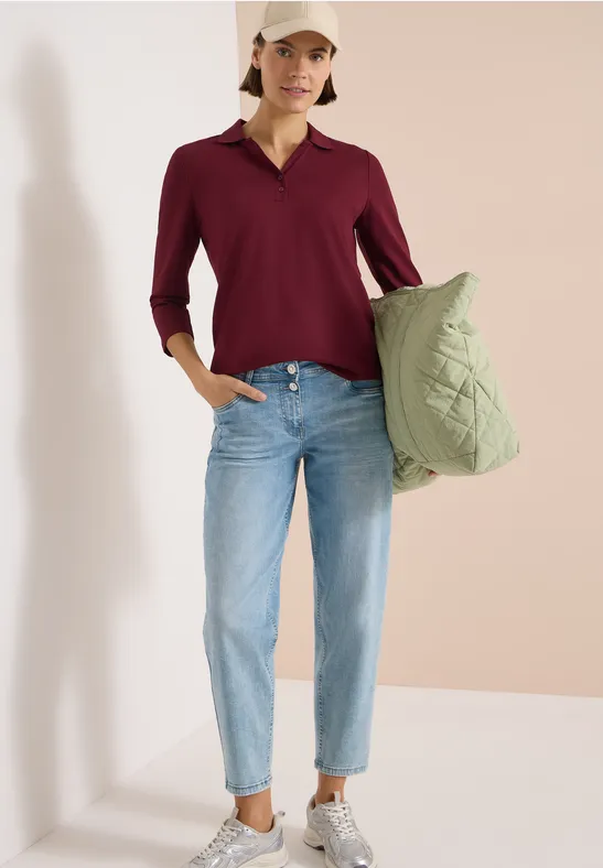 Frau trägt eine grüne Stepp-Tasche, bordeauxrote Bluse und helle Jeans vor hellem Hintergrund.