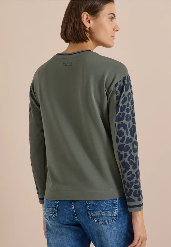 Frau von hinten in olivgrünem Langarmshirt mit animal-print-Ärmeln und Jeans vor hellem Hintergrund.