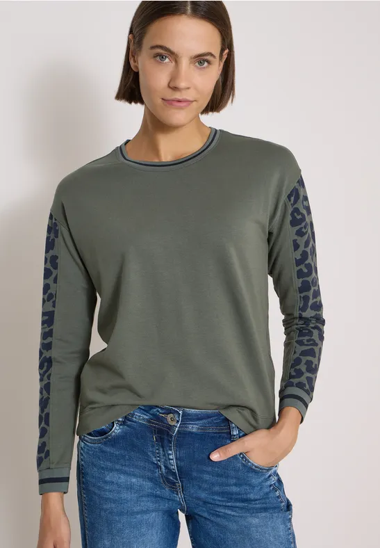 Frau mit braunen Haaren trägt olivgrünen Pullover mit gemusterter Passe an den Ärmeln und blaue Jeans.