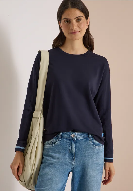 Vrouw in marineblauwe trui en jeans met beige schoudertas, voor een neutrale achtergrond.
