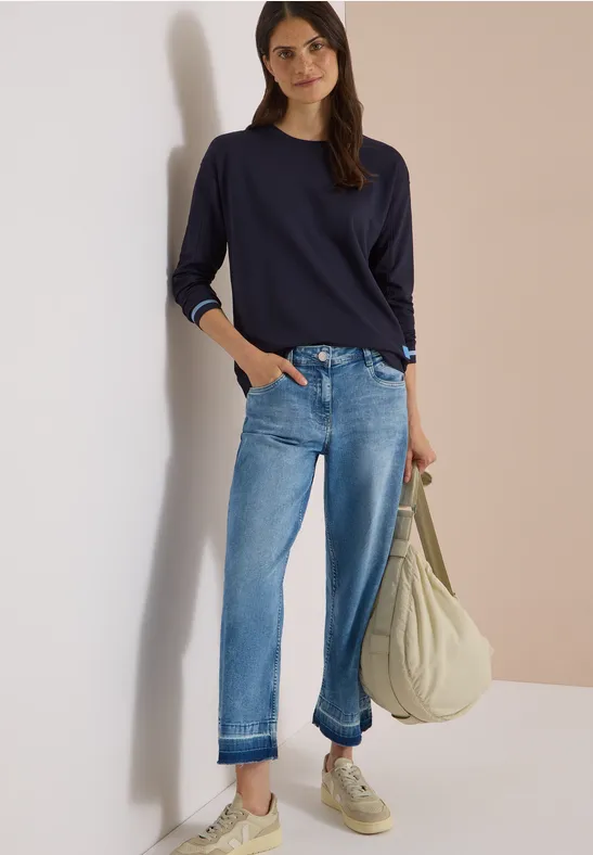 Vrouw met donkerblauw shirt, losse jeans en witte sneakers, glimlachend tegen muur, draagt grote beige tas.