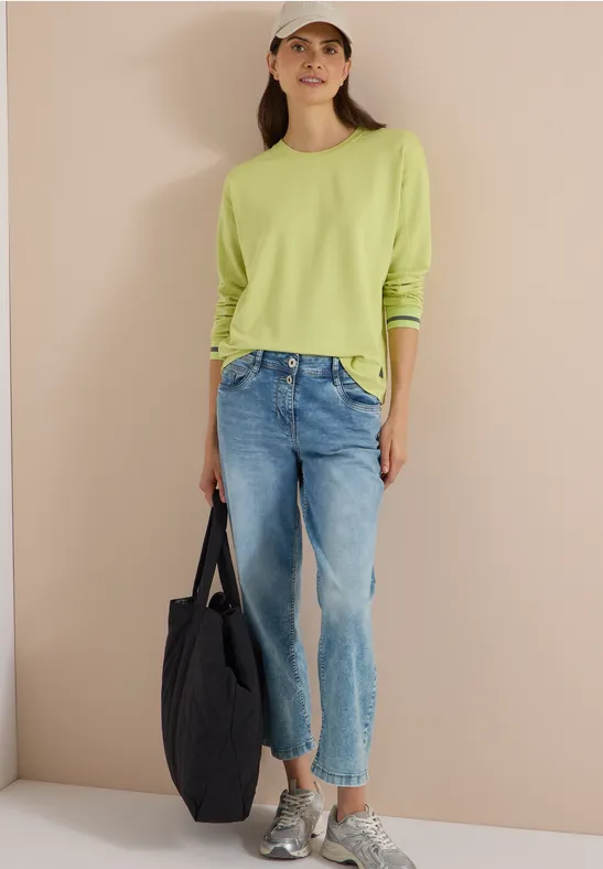 Vrouw draagt groen shirt, spijkerbroek, beige pet, sneakers, zwarte tas, lacht voor beige muur.