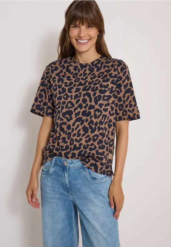Lächelnde Frau in Leo-T-Shirt und Jeans vor heller Wand.