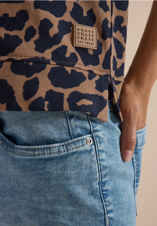 Nahaufnahme einer Person in hellblauen Jeans, Hand in Tasche, trägt ein Oberteil mit Leopardenmuster und Label.