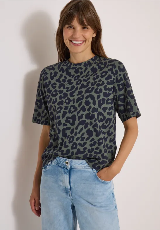 Frau in grünem Leo-T-Shirt und hellblauen Jeans, lächelt mit einer Hand in der Tasche.