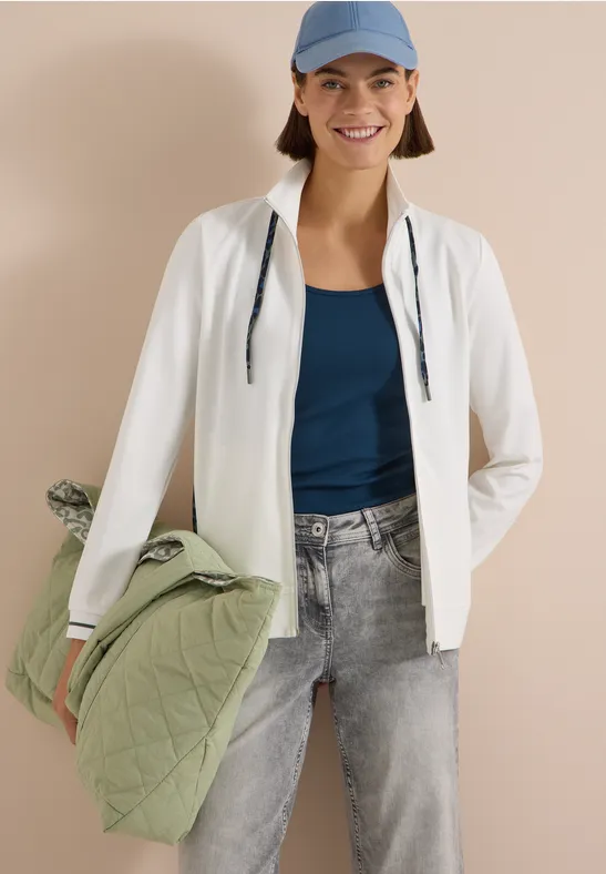 Femme souriante en veste blanche, débardeur bleu, jean et casquette, tenant une veste matelassée verte.