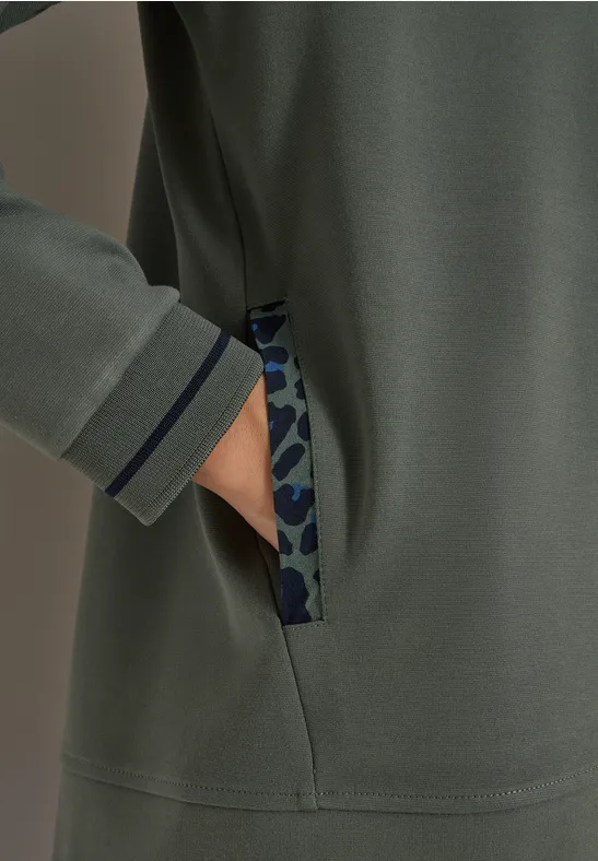 Hand in Seitentasche eines olivegrünen Sweatshirts mit camouflagefarbenem Besatz und dunkelblauem Saum.