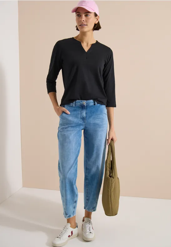 Frau in schwarzem 3/4-Arm-Shirt, hellblauen Jeans und weißen Sneakers, trägt eine olivgrüne Tasche.