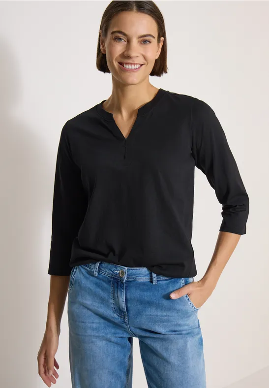 Frau in schwarzem Langarmshirt und blauen Jeans steht vor hellem Hintergrund.