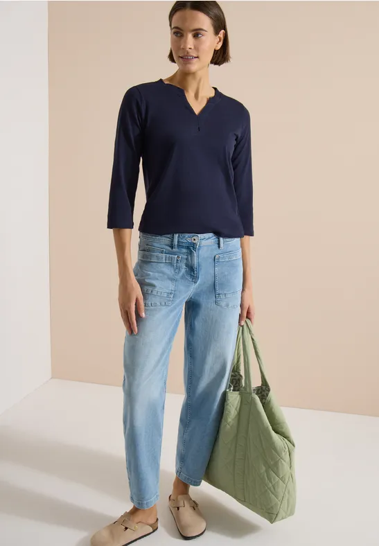 Frau in dunkelblauem V-Shirt, hellen Jeans, beigen Schuhen und grüner gesteppter Tasche vor neutralem Hintergrund.
