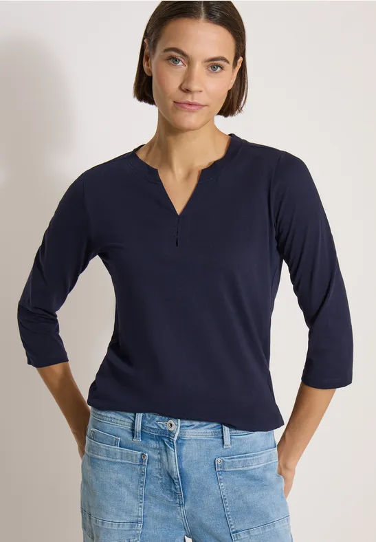 Dame trägt langärmliges dunkelblaues Shirt und helle Jeans, steht mit Händen in den Hüften.