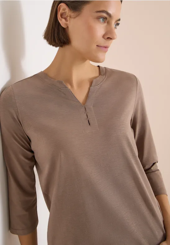 Eine Frau trägt ein taupefarbenes 3/4-Arm-Shirt mit V-Ausschnitt und Knopfleiste.