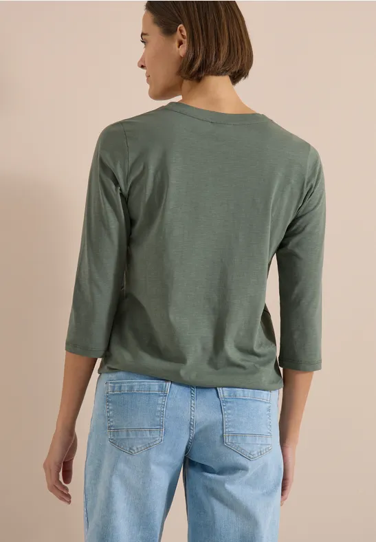 Rückansicht einer Person mit grünem 3/4-Arm-Shirt und blauen Jeans vor beigem Hintergrund