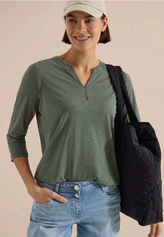 Eine lächelnde Frau in grüner 3/4-Arm-Bluse, Jeans und weißem Cap mit schwarzer Umhängetasche.