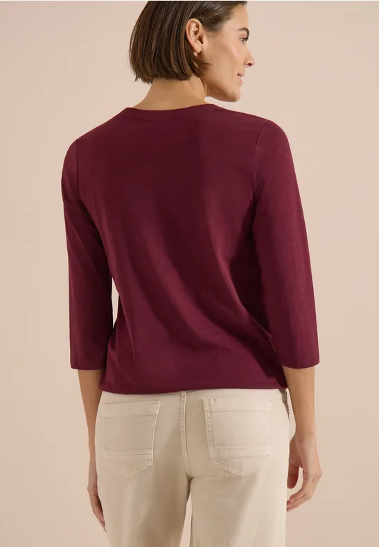 Rückansicht einer Frau in einem weinroten Pullover mit 3/4-Ärmeln und weißen Hosen.