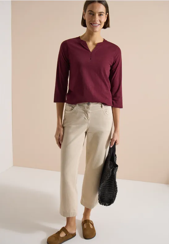 Frau in bordeauxrotem 3/4-Ärmel-V-Shirt, beigefarbenen Culottes, braunen Slippern und schwarzer Tasche.