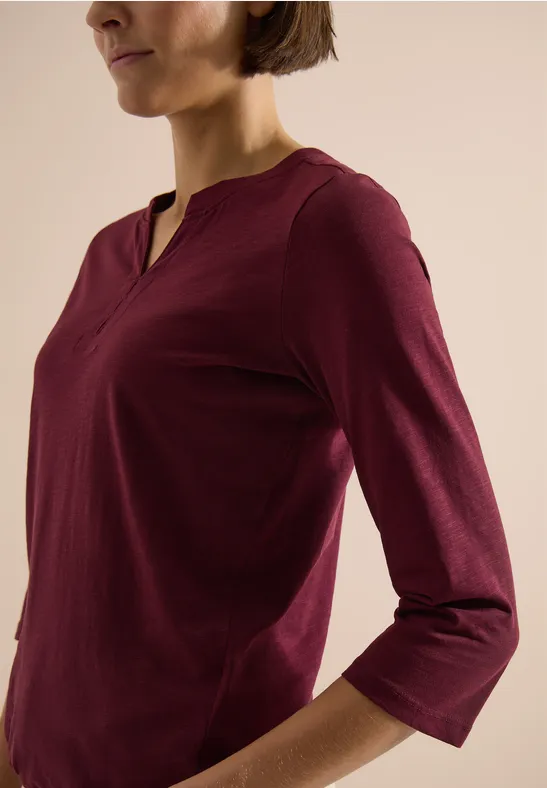 Oberkörper einer Person in burgunderrotem 3/4-Arm-Shirt vor hellem, neutralem Hintergrund.