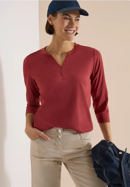 Frau trägt rotes Langarmshirt mit Knopfleiste und helle Hose, hält lässig eine Jacke in der Hand.
