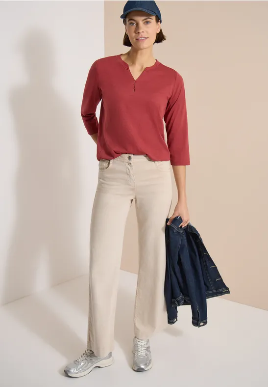 Person in rotem 3/4-Arm-Shirt, beigen Schlaghosen und silbernen Sneakern, hält eine Jeansjacke in der Hand.