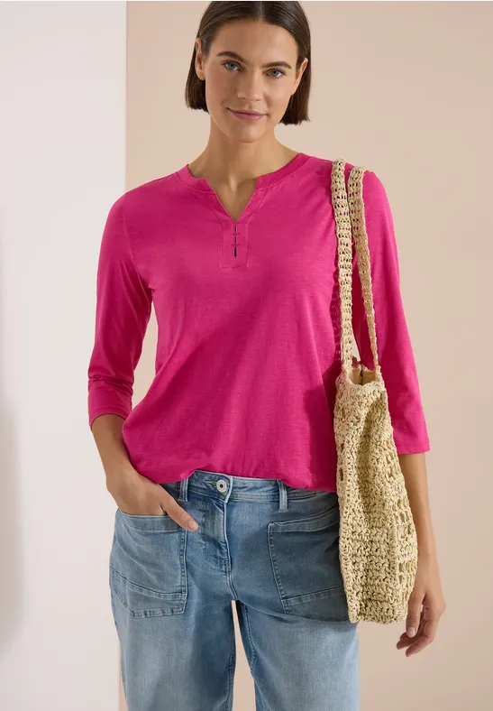 Frau trägt pinkes 3/4-Arm-Shirt, blaue Jeans und hält eine gehäkelte Strohtasche.